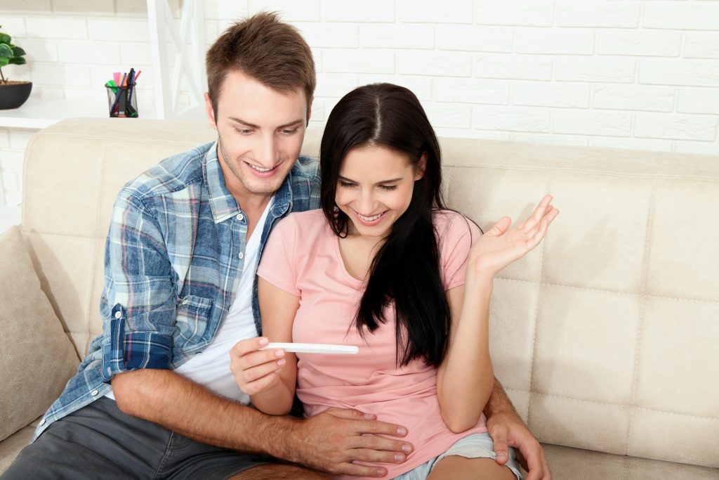 Šťastný párik s tehotenským testom v ruke v obývačke na sedačke.